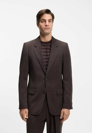 Mand stående iført en mørkebrun jakkesætjakke over en stribet mørkebrun sweater mod en ensfarvet hvid baggrund.