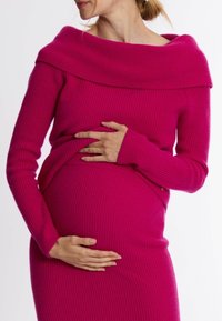 Rippenshirt mit schulterfreiem Design in leuchtendem Pink, taillierte Passform, lange Ärmel, weiche Textur, über einem passenden gerippten Kleid getragen, Hände auf dem Bauch.