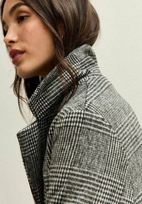 Abrigo a cuadros grises y negros con un alto cuello, hecho de un tejido texturizado. El diseño presenta líneas claras y una silueta estructurada.