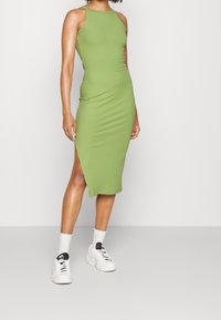 Nainen, jolla on ylleen pukeutunut hihaton, ribattu, vaaleanvihreä midi-mekko, jossa on sivuhaalari, valkoiset sukat ja valkoiset lenkkarit mustilla yksityiskohdilla.