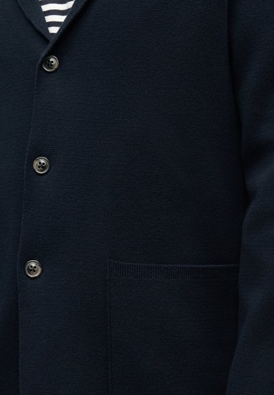 Cardigan bleu marine foncé avec trois boutons ronds et une petite poche avant, porté par-dessus une chemise rayée noire et blanche.