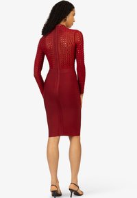 Rotes, figurbetontes Kleid mit langen Mesh-Ärmeln und Nietenverzierung, hohem Ausschnitt und verdeckt eingearbeitetem Reißverschluss auf der Rückseite. Strukturierter Stoff mit glatter Oberfläche.