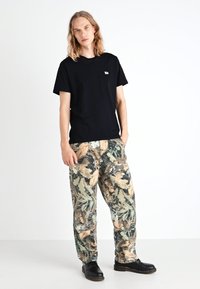 T-shirt nera in cotone con un piccolo logo, abbinata a pantaloni cargo larghi in tonalità beige e verde militare. Le scarpe nere completano il look.
