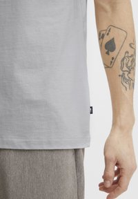 Grijze katoenen t-shirt met een gladde textuur, iets losse pasvorm en een klein merklabel aan de linkerkant. Zichtbare arm tatoeage en tan broek.