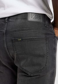 Vaqueros de denim negro con un parche de cuero de la marca Lee en la cintura, que presentan un diseño clásico de bolsillo trasero y detalles de costura sutiles.