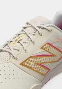 Gros plan sur une basket beige avec des lacets blancs et un logo "N" doré et rose sur le côté, présentant des détails perforés et une languette en mesh.