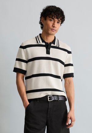 Jeune homme aux cheveux bouclés porte un polo blanc à manches courtes en tricot avec des rayures horizontales noires et un pantalon noir avec ceinture, debout contre un mur gris.