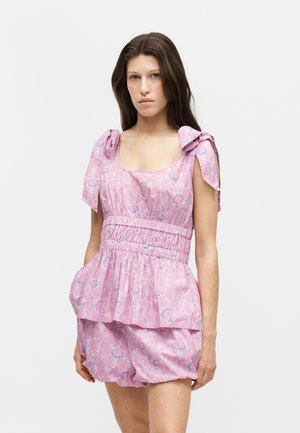 Vrouw draagt een roze bloemenmouwloze jurk met strikjes op de schouders, geplooide taille, peplumdetaillering en bijpassende shorts tegen een effen achtergrond.