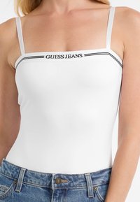 Biely body s tenkými ramienkami, zdobené čiernym logom "GUESS JEANS" a horizontálnymi čiernymi akcentmi na výstrihu. Nosené s modrými džínsovými šortkami.
