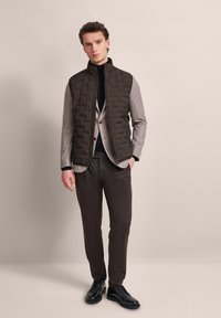 Padded dunkelgrüne Weste über einem hellgrauen Blazer, kombiniert mit braunen Hosen und schwarzen Business-Schuhen. Glatte Texturen und taillierte Passform.