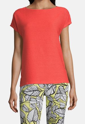 Vrouw draagt een gestructureerde rood top met korte mouwen en lichtgroene broek met grote zwart-witte bladprints.