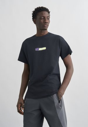 Fiatal férfi fekete pólóban, amelyen kétszínű, sárga és lila Nike logó látható, egy kézzel a zsebében áll, egyszínű háttér előtt.