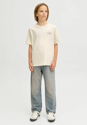 Maglietta in cotone beige a maniche corte e collo rotondo, con un piccolo logo stampato sul petto, abbinata a jeans azzurri chiari e sneakers.