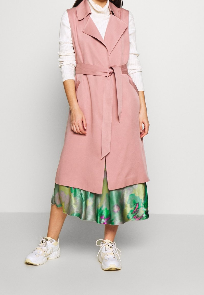 Růžový bezrukávový trenčkot s páskem, s klopovým límcem a bočními kapsami, nošený přes vzdušnou zelenou květinovou sukni.