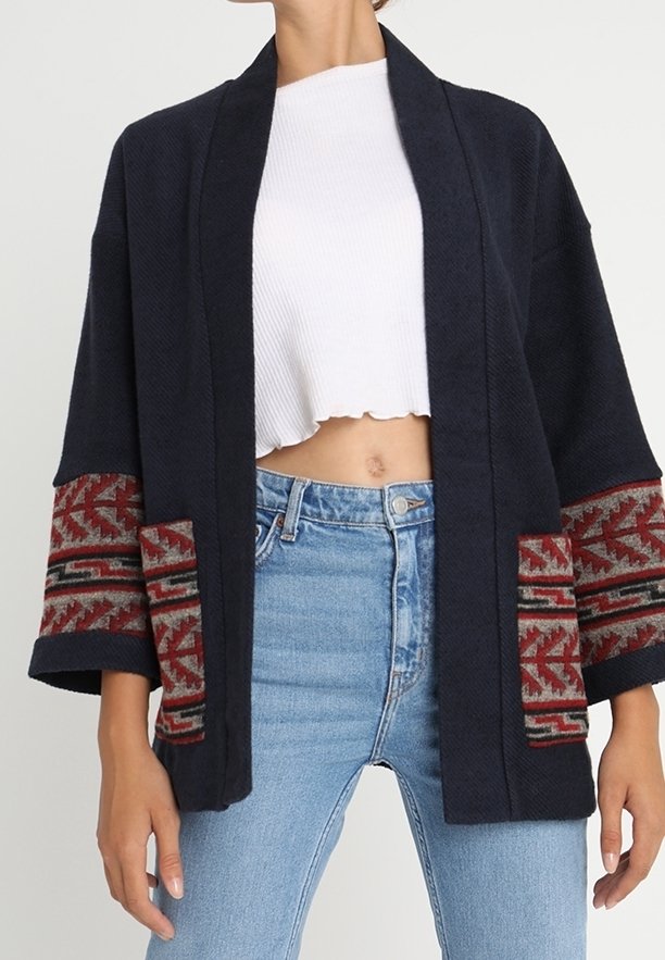 Femme portant un cardigan bleu marine ouvert avec des bords à motifs rouges sur les manches et les poches, un haut blanc court et un jean bleu clair.