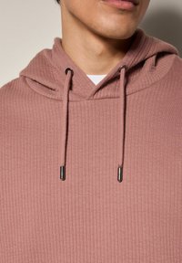 Sudadera de color rosa con canalé y capucha ajustable, con puntas metálicas plateadas en los cordones. La tela tiene una textura suave.