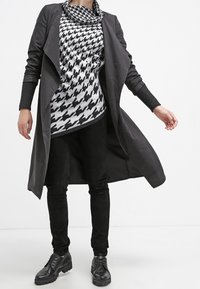 Czarny długi płaszcz o zwiewnym kroju, nałożony na czarno-biały top w wzór pepitki. Połączony z czarnymi spodniami i błyszczącymi czarnymi butami.