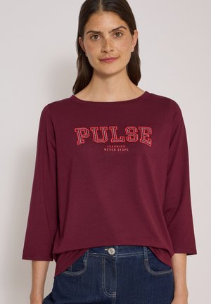Femme aux longs cheveux bruns portant une chemise bordeaux avec les textes "PULSE" et "LEARNING NEVER STOPS", associée à un jean bleu.