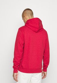 Sudadera roja de tejido suave y texturizado con una capucha amplia y puños acanalados. Cuenta con un bolsillo tipo canguro en la parte frontal y un corte relajado.