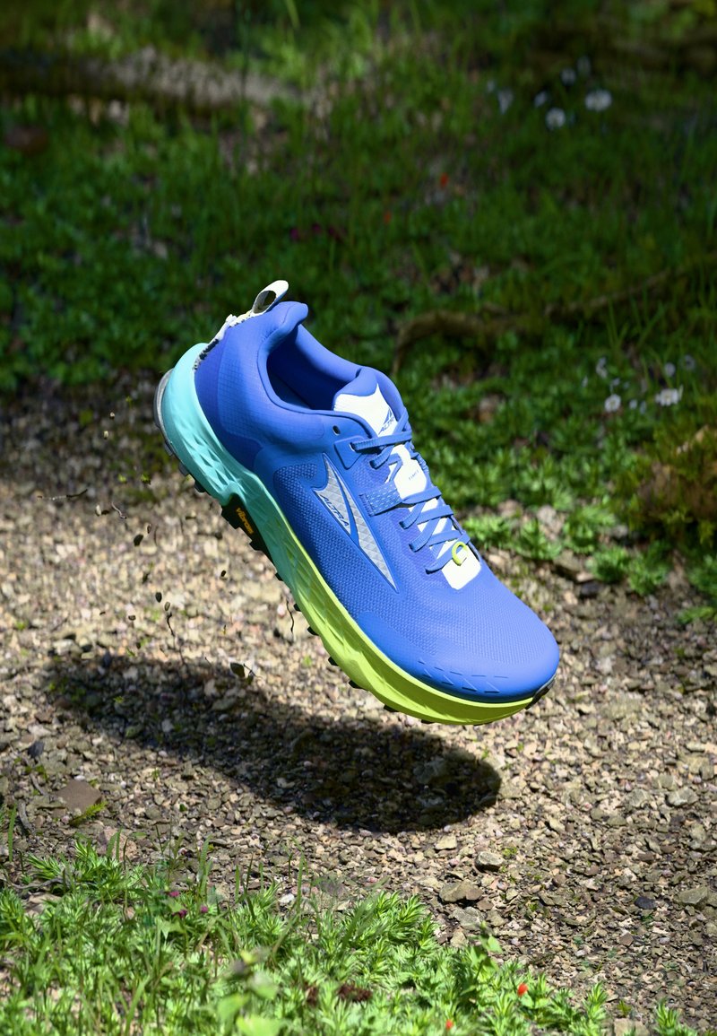 Blue running shoe with a textured upper, white accents, and a green sole, shown mid-air over a gravel path and grassy background.