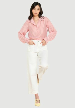 Naine poseerib käed taskutes, seljas roosa triibuline pluus, valged kõrge vöökohaga püksid ja kollased jalatsid ühevärvilise taustaga.