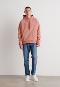 Helle rosa Kapuzenjacke mit Fronttasche und Kordelzug, mit einem strukturierten Logo. Kombiniert mit blauen Jeans und weißen Sneakern auf einer grauen Oberfläche.