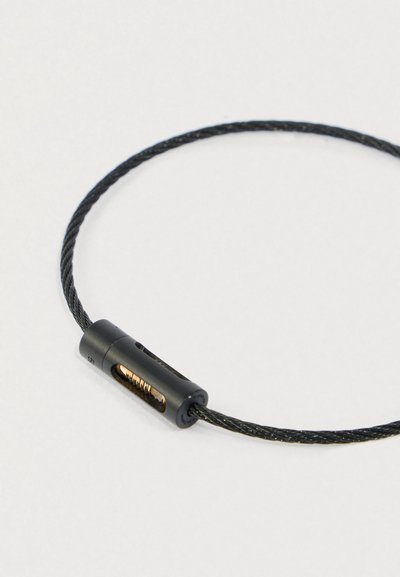 Melnā pītā tērauda troses cilpa ar mazu cilindriskā slēdzenes mehānismu, kurā iekšā redzams zelta atsperes.