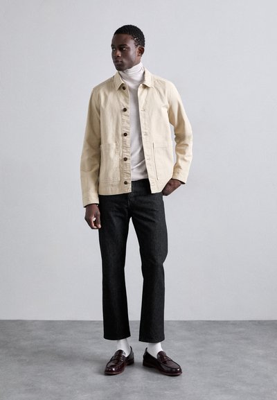 Chaqueta de algodón beige con cierre de botón, combinada con un suéter de cuello alto blanco, pantalones de mezclilla oscuros y mocasines marrones, sobre un fondo gris.