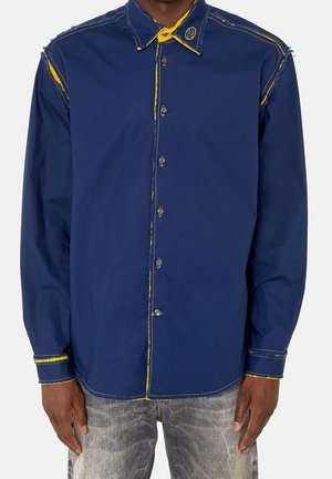 Homme portant une chemise bleue à boutons avec des coutures jaunes effilochées, associée à un jean gris délavé.