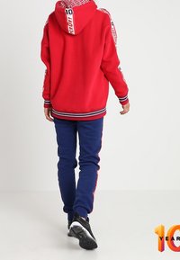Röd hooded sweatshirt med logotypdetaljer och marinblå byxor. Har ribbstickade muddar och fåll, kombinerat med svarta sportskor.