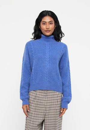 Helles blaues Strickpullover mit hohem Kragen, Kabelmustern und gerippten Bündchen, getragen über karierter Hose. Weiche Textur, entspannte Passform.