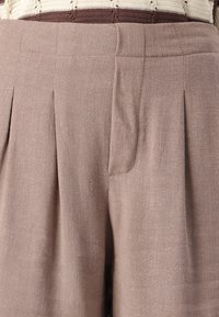 Pantalons en lin marron clair avec des plis, une texture lisse et une taille ajustée. Poche latérale visible intégrée dans le design.