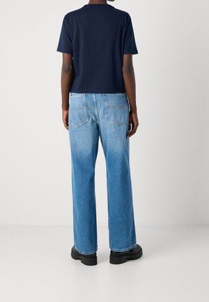 Person, der står med ryggen til iført en marineblå kortærmet skjorte, lyseblå lige jeans og sorte chunky sko på et hvidt gulv.