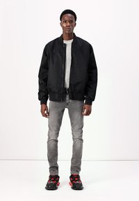 Chaqueta bomber negra con cremallera frontal, cuello y puños acanalados; combinada con jeans grises y zapatos negros con acentos rojos.