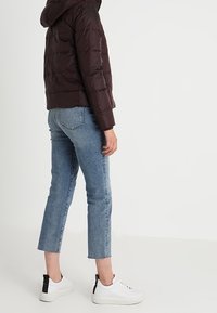 Veste matelassée marron foncé avec capuche, jean bleu court, et baskets blanches avec accent talon noir. Vue de côté, arrière-plan minimal.