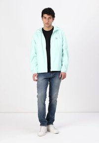 Licht mintgroen waterdicht jack met een capuchon, voorkant rits en logo; gecombineerd met een zwart shirt, blauwe spijkerbroek en witte sneakers.