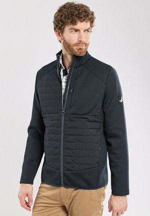 Veste bleu marine pour homme avec fermeture éclair sur le devant, panneau frontal matelassé, dos texturé et poches latérales. Dispose d'un col montant et d'un accent logo subtil.