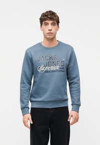 Νεαρός άνδρας με σγουρά μαλλιά, που φοράει μπλε φούτερ με το κείμενο "Jack & Jones Superior New Aesthetics", στέκεται μπροστά σε απλό λευκό φόντο.