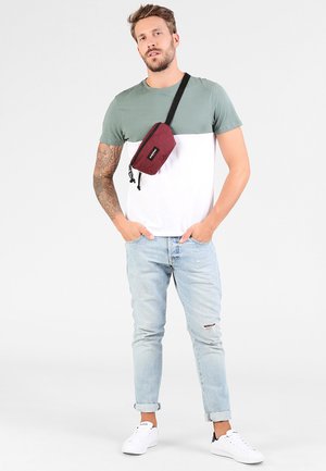T-shirt a blocchi di colore verde e bianco con maniche corte, abbinato a jeans strappati blu chiaro e sneakers bianche. Si indossa una borsa a tracolla bordeaux.