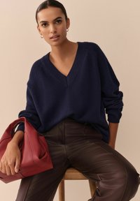 Navy V-Ausschnitt-Pullover mit überschnittenen Schultern, kombiniert mit braunen Lederhosen. Hält eine strukturierte rote Handtasche und sitzt auf einem Hocker aus Holz.