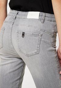 Jeans de mezclilla gris con un diseño ajustado, cintura de altura media, bolsillos traseros con un detalle metálico y una etiqueta de marca blanca en la cintura.