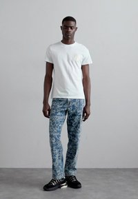 Λευκό κοντομάνικο μπλουζάκι με λογότυπο στο αριστερό στήθος, συνδυασμένο με μπλε παντελόνια με σχέδια που περιλαμβάνουν αφηρημένα γραffiti. Μαύρα αθλητικά παπούτσια.