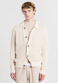 Cardigan lavorato a maglia di un beige chiaro con un motivo testurizzato, bottoni grandi e un colletto, abbinato a una maglietta crema sotto.