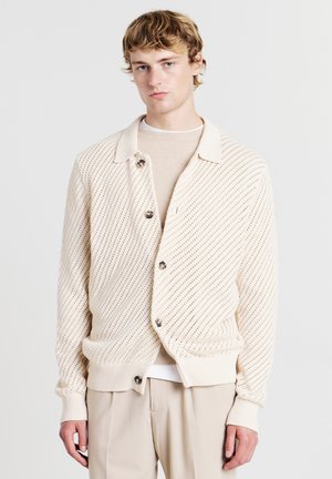 Cardigan lavorato a maglia di un beige chiaro con un motivo testurizzato, bottoni grandi e un colletto, abbinato a una maglietta crema sotto.