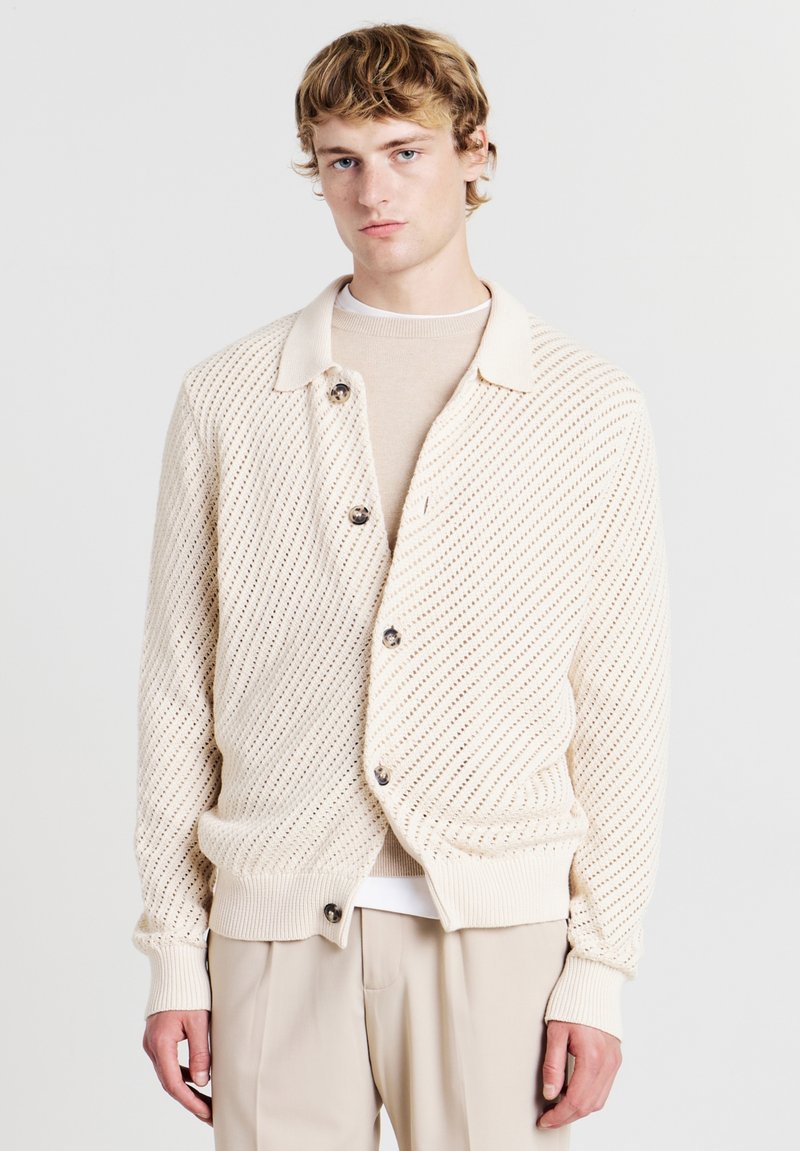 Helle beige Strickjacke mit einem strukturierten Muster, großen Knöpfen und einem Kragen, kombiniert mit einem cremigen Oberteil darunter.