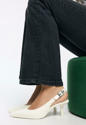 Pantofi cu toc alb cu bareta în spate și vârf ascuțit, având un detaliu cu cataramă argintie. Purtate cu blugi evazați închiși la culoare, prezentând o textură netedă.