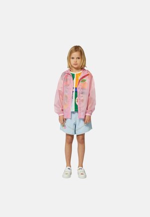 Giacca rosa traslucida con cappuccio, indossata sopra una maglietta grafica colorata e pantaloni corti azzurri chiari con toppe. Scarpe bianche senza lacci.