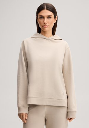 Donna con capelli castani lisci che indossa una felpa beige con cappuccio e pantaloni abbinati, in piedi davanti a uno sfondo chiaro e uniforme.