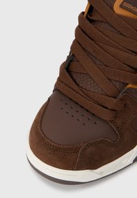 Brown sneaker featuring a suede and leather upper, textured brown laces, perforated toe cap, and white midsole with rubber outsole.