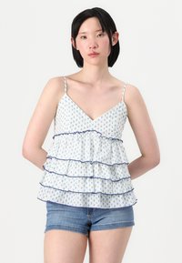 Top blanco con estampado floral, con volantes en capas, detalles azules y tirantes ajustables, combinado con pantalones cortos de mezclilla.